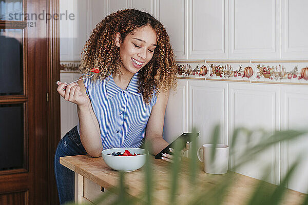 Schöne Frau  die ein Mobiltelefon benutzt  während sie in der heimischen Küche einen Obstsalat isst