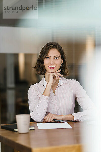 Schöne junge Geschäftsfrau sitzt mit Hand am Kinn am Schreibtisch im Büro