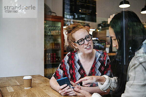 Frau mit Brille im Gespräch mit einer Freundin  die ein Mobiltelefon in einem Café benutzt