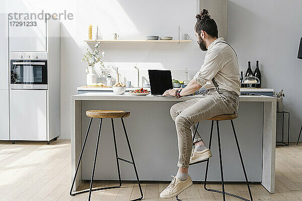 Mittlerer erwachsener Mann  der einen Laptop benutzt  während er zu Hause in der Küche sitzt