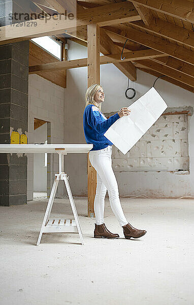 Lächelnde Bauunternehmerin mit Bauplan  die wegschaut und sich auf einen Tisch auf der Baustelle stützt