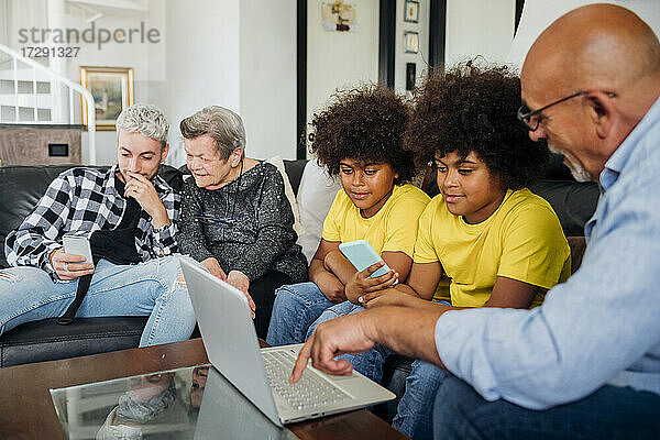 Zwillingsjungen mit Großvater  der einen Laptop benutzt  während ein junger Mann mit einem Smartphone bei der Urgroßmutter im Wohnzimmer sitzt