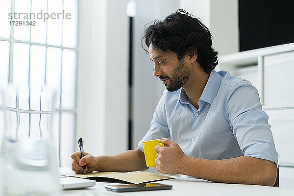 Männlicher Unternehmer mit Kaffeetasse  der im Büro in ein Buch schreibt