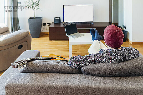 Männlicher Gitarrist beim Fernsehen auf dem Sofa zu Hause