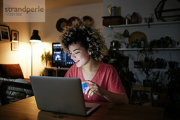 Lächelnde junge Frau sitzt mit Laptop und Kreditkarte zu Hause