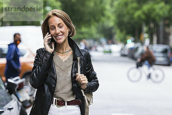 Lächelnde Frau mit Umhängetasche  die auf einer Straße in der Stadt mit einem Mobiltelefon spricht