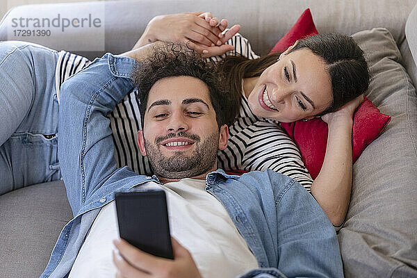 Glückliches Paar  das ein Mobiltelefon benutzt  während es zu Hause auf dem Sofa liegt