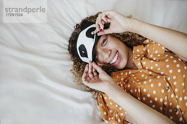 Lächelnde junge Frau mit Augenmaske  während sie zu Hause auf dem Bett liegt