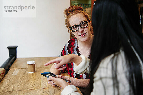 Frau mit Mobiltelefon  die eine Freundin ansieht  die im Café gestikuliert