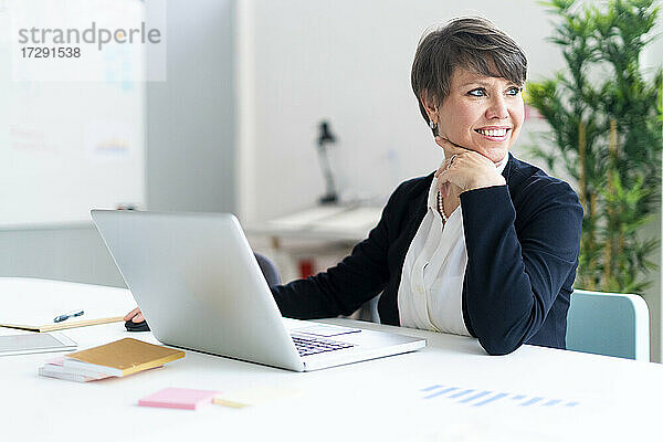 Lächelnde weibliche Fachkraft sitzt mit Laptop im Büro und schaut weg