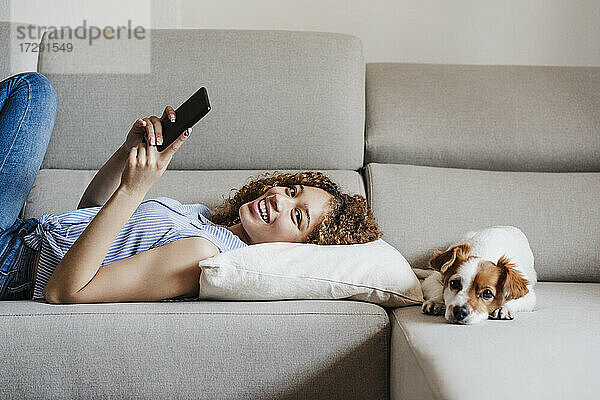 Lächelnde Frau mit Handy  die neben einem Jack Russell Terrier auf dem Sofa zu Hause liegt