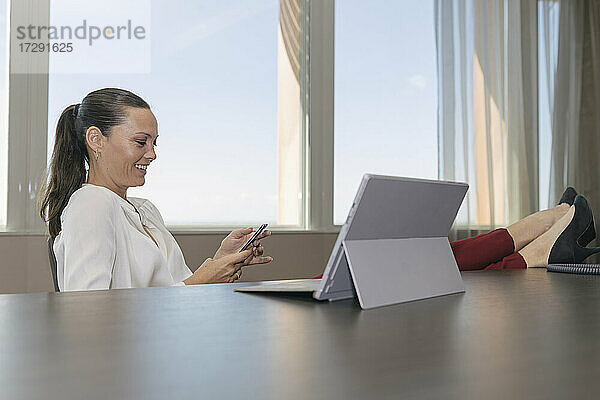 Lächelnde Geschäftsfrau  die über ihr Smartphone Textnachrichten verschickt  während sie am Schreibtisch im Büro sitzt