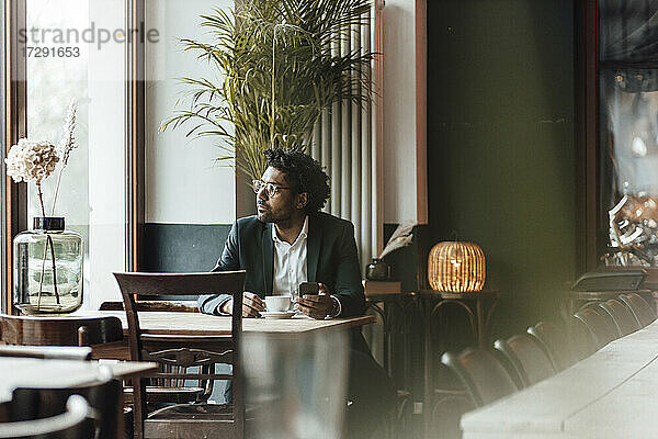 Älterer Geschäftsmann mit Mobiltelefon sitzt mit Kaffee im Café und schaut weg