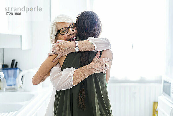 Lächelnde ältere Frau mit geschlossenen Augen  die ihre Enkelin in der Küche zu Hause umarmt