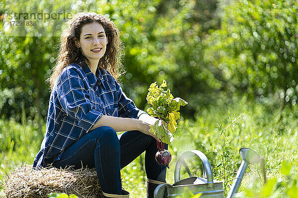 Lächelnde junge Frau sitzt auf einem Stapel Heu und hält eine Rote-Bete-Pflanze