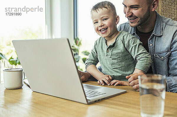 Fröhlicher Sohn und Vater sitzen mit Laptop und führen einen Videoanruf zu Hause durch