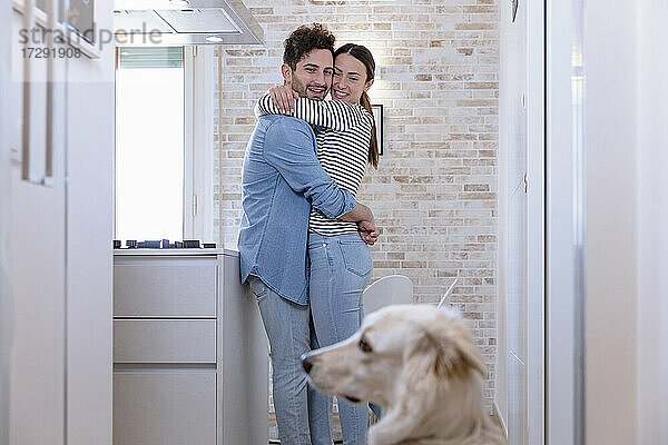 Lächelndes  sich umarmendes Paar mit Blick auf den Hund in der heimischen Küche zu Hause