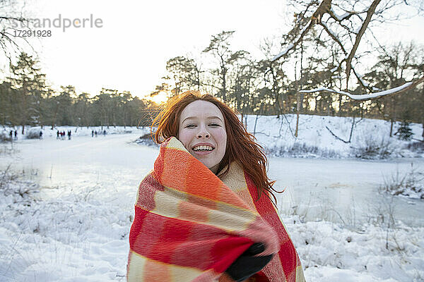 Fröhliche rothaarige Frau in Decke eingewickelt im Winter bei Sonnenuntergang