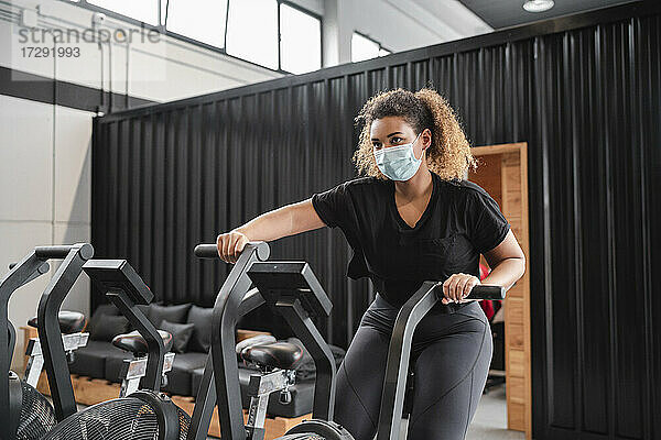Aktive Frau mit Schutzmaske beim Radfahren im Fitnessstudio während COVID-19
