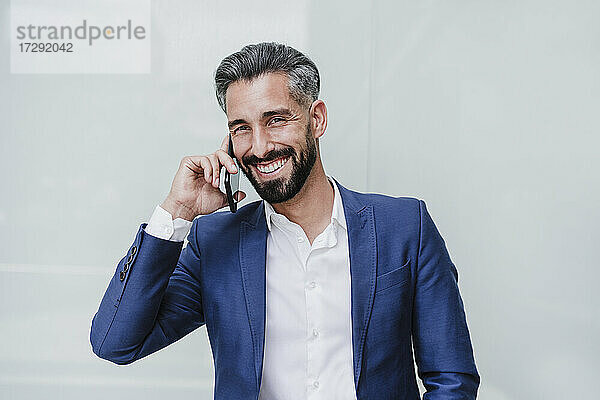Männlicher Unternehmer lächelt  während er vor einer Wand mit seinem Handy spricht