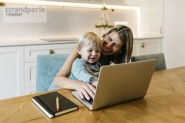 Lächelnder Junge mit Laptop und Mutter  die zu Hause arbeitet