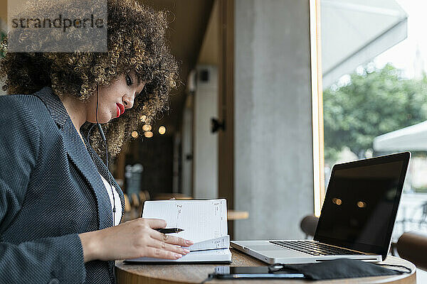 Berufstätige Frau mit Kopfhörern  die in einem Café sitzend in einem Tagebuch sucht