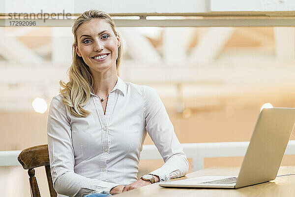Lächelnde Unternehmerin mit Laptop am Schreibtisch in einem kreativen Büro
