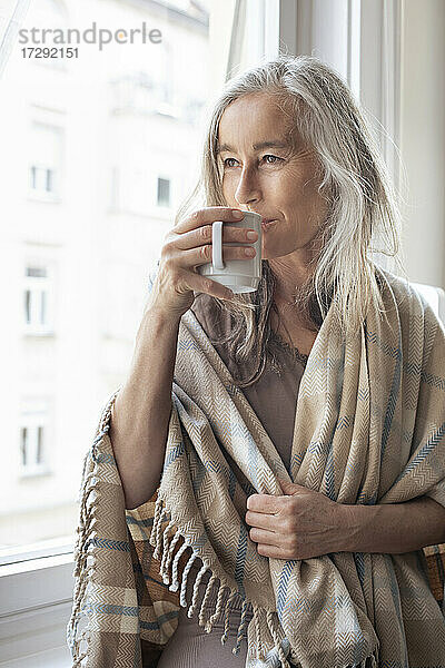Reife Frau mit weißen Haaren schaut durch das Fenster und trinkt zu Hause Kaffee