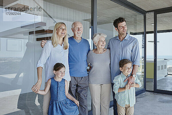 Lächelnde Mehrgenerationenfamilie  die gemeinsam vor einer Glaswand steht und wegschaut