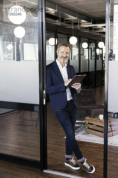 Lächelnder männlicher Unternehmer  der einen Laptop hält und sich an eine Glastür im Büro lehnt