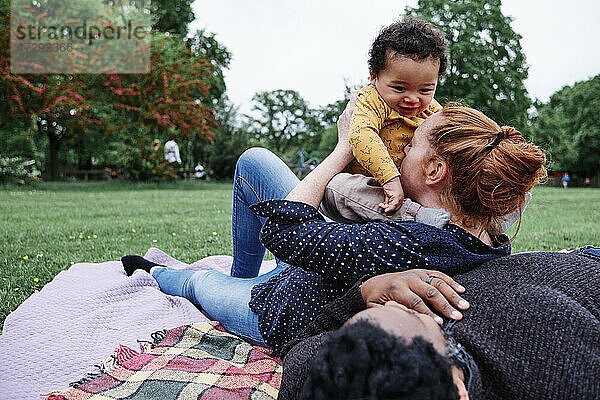 Mutter küsst ihren Sohn und lehnt sich an einen Mann  der auf einer Picknickdecke im Park liegt