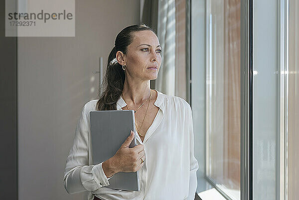 Unternehmerin mit digitalem Tablet  die durch ein Fenster schaut  während sie im Büro steht