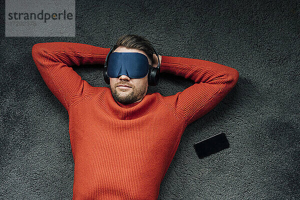 Männlicher Berufstätiger  der mit einer Augenmaske ein Nickerchen macht  während er am Arbeitsplatz über Kopfhörer Musik hört