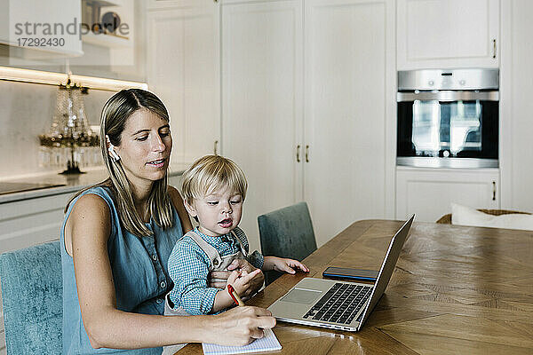 Mittlere erwachsene Frau  die während eines Videoanrufs in ihr Tagebuch schreibt  während ihr Sohn zu Hause auf dem Schoß sitzt