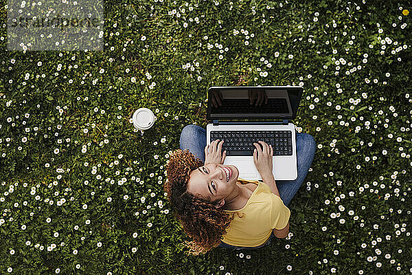 Junge Frau  die mit ihrem Laptop auf einer Wiese inmitten von Blumen sitzt und nach oben schaut