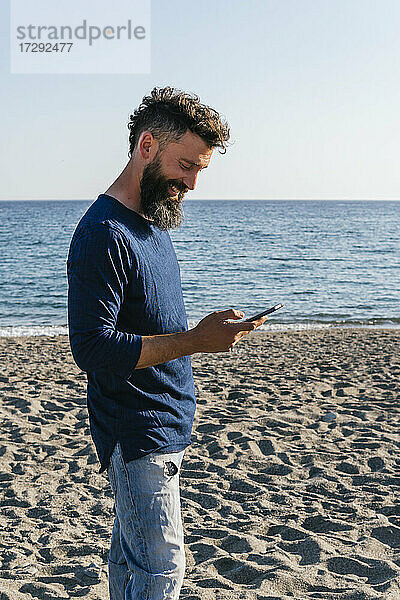 Lächelnder Mann  der am Strand stehend ein Mobiltelefon benutzt