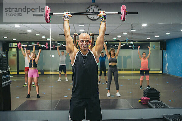 Lächelnder männlicher Ausbilder  der eine Langhantel hebt  während er mit Männern und Frauen im Fitnessstudio trainiert
