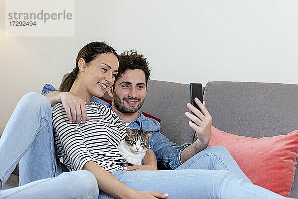 Lächelndes Paar mit Katze nimmt Selfie durch Smartphone zu Hause