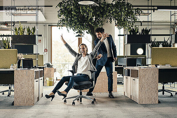 Fröhliche Geschäftsfrau auf einem Stuhl sitzend mit einem im Hintergrund stehenden Kollegen im Büro