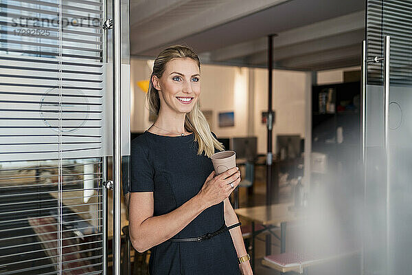 Lächelnde junge Geschäftsfrau mit wiederverwendbarem Kaffeebecher  die sich an eine Glastür lehnt und wegschaut