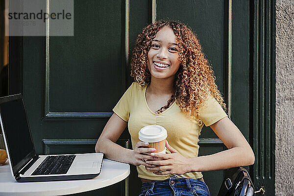 Lächelnde junge Frau mit Laptop  die einen wiederverwendbaren Becher hält  während sie in einem Straßencafé sitzt