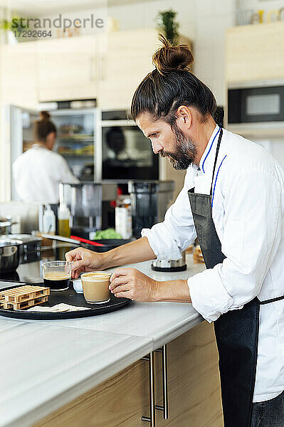 Männlicher Koch ordnet Kaffeetassen auf einem Tablett in einer Großküche an