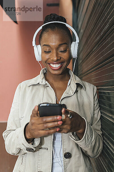 Glückliche Frau mit Kopfhörern  die eine Textnachricht über ihr Mobiltelefon sendet  während sie Musik hört  die von einem Fensterladen kommt