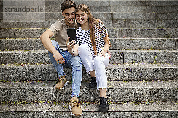 Lächelndes Paar  das ein Selfie mit dem Mobiltelefon macht  während es auf den Stufen sitzt
