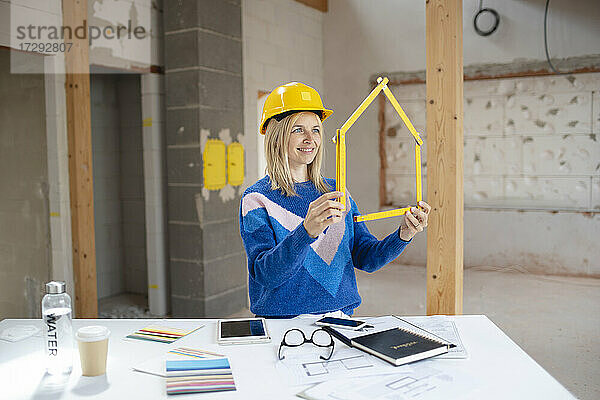 Lächelnde Bauunternehmerin  die auf einer Baustelle mit einem Zollstock ein Haus formt