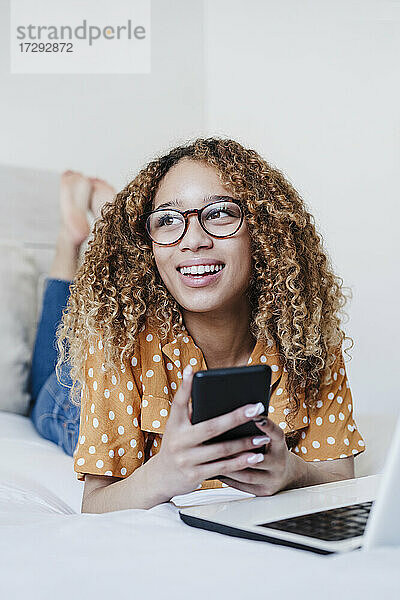 Junge Frau mit Mobiltelefon  die wegschaut  während sie zu Hause auf dem Bett liegt