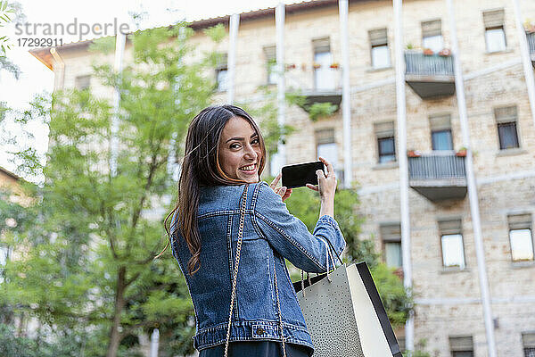 Lächelnder weiblicher Tourist mit Einkaufstaschen  der ein Selfie mit seinem Smartphone in der Stadt macht