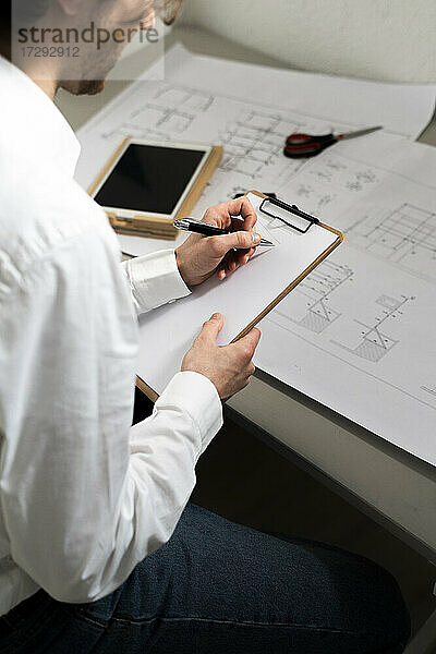 Männlicher Grafikdesigner schreibt in ein Klemmbrett  während er am Schreibtisch im Büro sitzt