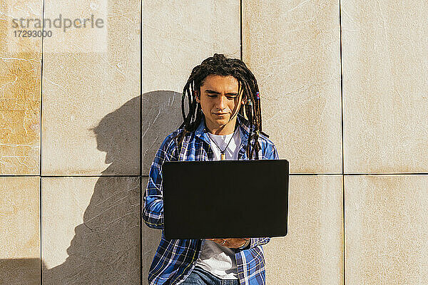 Junger Mann mit Dreadlocks benutzt Laptop und lehnt sich an die Wand