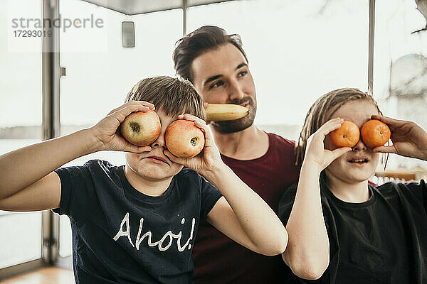 Verspielte Brüder bedecken ihre Augen mit Früchten  während sie mit ihrem Vater in der Küche sitzen